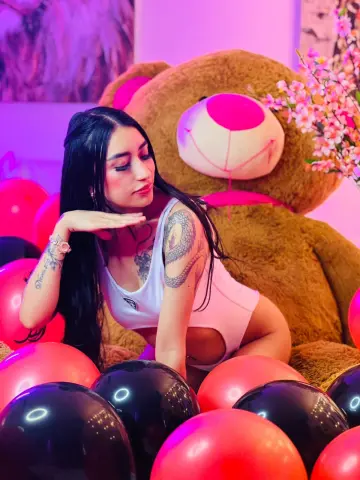 Tattooed woman in a white bodysuit sitting among red and black balloons by a giant teddy bear in pink lighting.