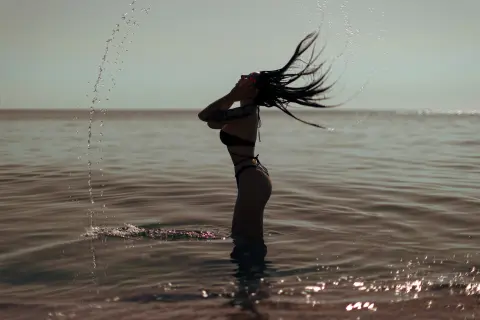 Silhouette of a woman in a black bikini splashing water in the ocean during a dreamy sunset.