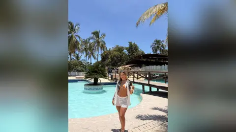 Young woman in a white summer outfit relaxing by a tropical resort swimming pool under a sunny blue sky.