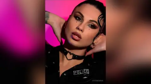 CutemEmma poses with her hands behind her head, wearing a black police-style outfit and a heart-shaped choker against a pink background.