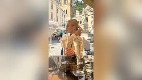 AmaFox — Stylish woman in a white blouse enjoying champagne at an outdoor European cafe on a sunny summer day.