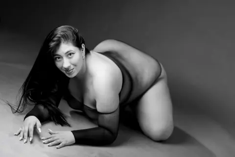 Black and white portrait of a curvy woman in sheer black lingerie posing on a studio floor with a moody vibe.