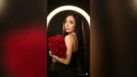 Elegant woman in a black dress holding a large bouquet of red roses against a glowing ring light background.