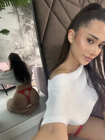 Young woman in a white ribbed crop top and red bikini bottoms posing in a modern bedroom setting.
