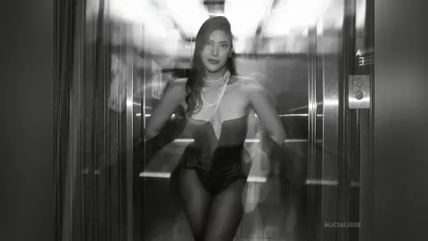 Elegant woman in a black bodysuit and pearl necklace posing inside a modern elevator in a moody black and white style.