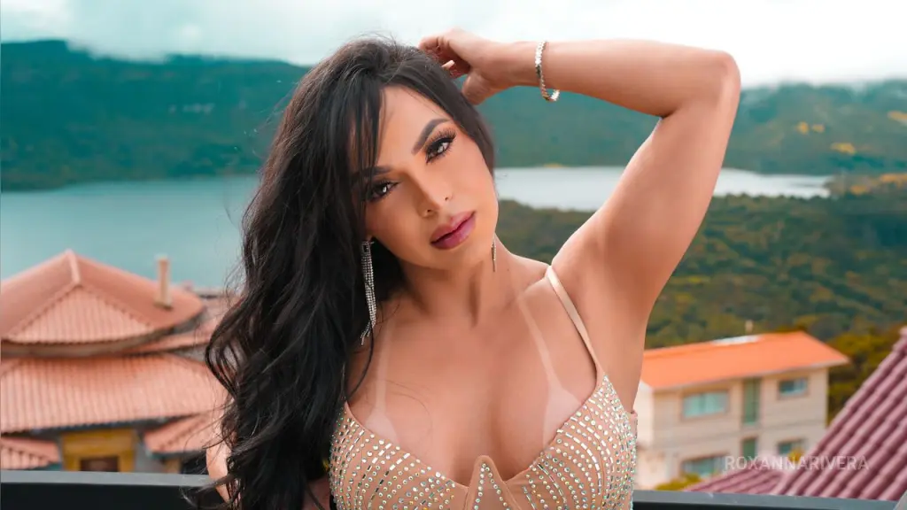 Roxanna — RoxannaRivera poses with her hand in her long dark hair, wearing a sparkly tan bra against a scenic mountain and lake background.