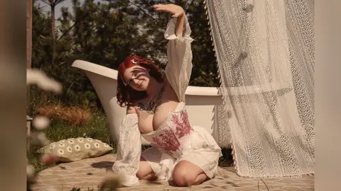 Redheaded woman in a white lace corset outfit posing outdoors in a dreamy garden setting.