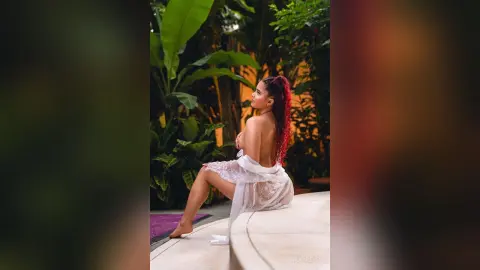 Red-haired woman in a white lace cover-up sitting by a tropical garden pool in a serene mood.