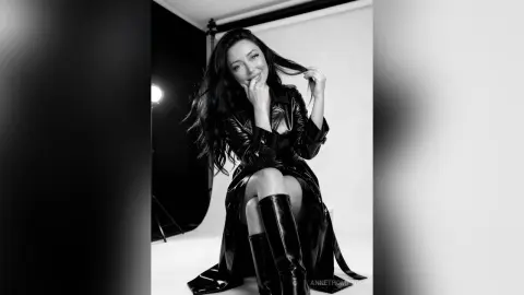 Smiling woman in a black leather trench coat and tall boots posing in a studio setting.