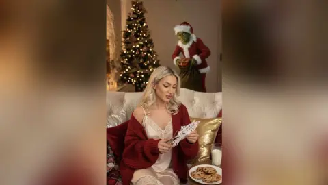 JessycaPage sits on a white couch in a red cardigan and lace camisole, holding paper snowflakes near a decorated Christmas tree.