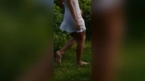 A woman in a white summer dress walking barefoot through a lush green garden in soft natural light.
