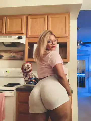 Curvy woman in pink crop top and grey lounge shorts posing in a cozy kitchen setting.
