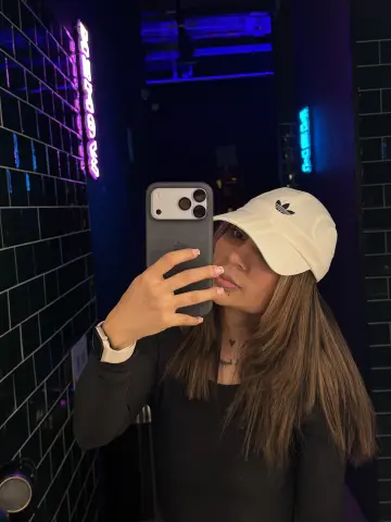 Young woman in a black long-sleeve shirt and white Adidas cap taking a selfie in a moody neon-lit room.