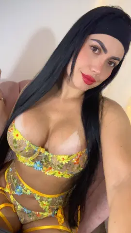 Smiling brunette woman in yellow floral lace lingerie sitting in a bright indoor setting.