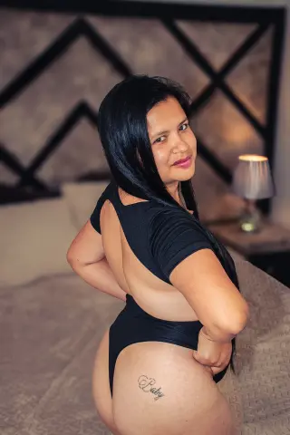 Smiling woman in a black bodysuit posing on a bed in a cozy bedroom setting.