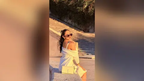 Stylish woman in a white summer dress and sunglasses sitting on stone stairs during a golden hour sunset.