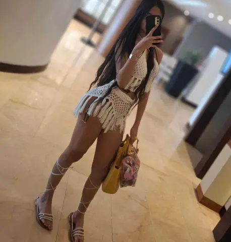 Woman in white crochet mini dress and strappy sandals posing in a modern luxury hotel lobby.