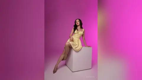 Elegant woman in a pale yellow summer dress sitting on a white pedestal against a vibrant pink background.