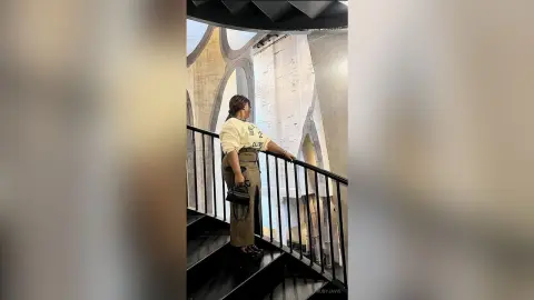 Woman in cream sweatshirt and olive trousers standing on a modern staircase in a minimalist architectural setting.