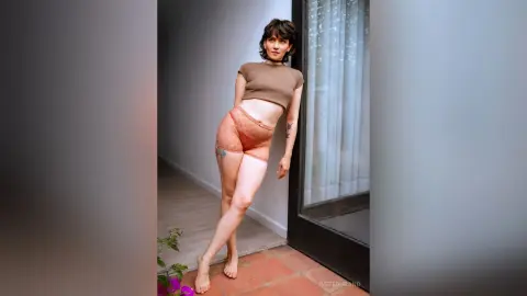 IveteDurand poses in a brown crop top and peach lace panties, leaning against a glass door in a bright room.