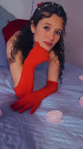 Woman in red evening gloves and headband relaxing on a bed with pink rose petals in a romantic mood.
