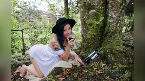 Tattooed woman in a white dress and black hat sipping red wine in a lush, moody forest setting.