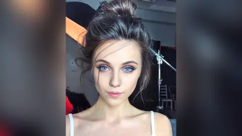 Young woman with messy bun and soft makeup looking into camera in a studio setting.