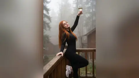 Redheaded woman in black athletic wear holding a wine glass on a misty forest balcony.