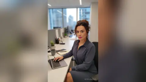 Professional woman in a grey blouse working on a laptop at a modern office desk with a city view.