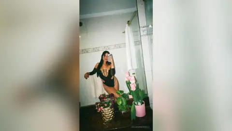 Woman in a black cutout bodysuit taking a mirror selfie in a bright bathroom with pink flowers.