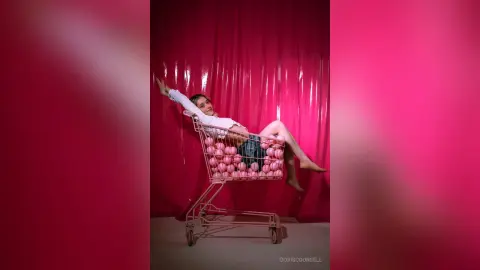 Young woman in white shirt and denim shorts reclining playfully in a pink shopping cart against a pink backdrop.