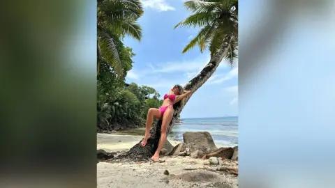 Katia poses in a pink bikini while leaning against a palm tree on a tropical beach.