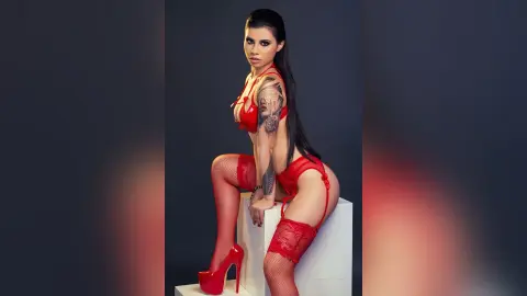 Tattooed woman in red lace lingerie and stockings posing confidently in a studio setting.