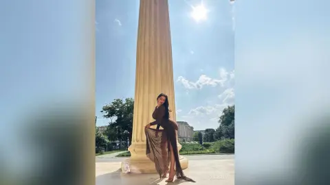 NicoleBella stands outdoors next to a tall stone column, wearing a long brown dress with a high slit that shows her bare leg.