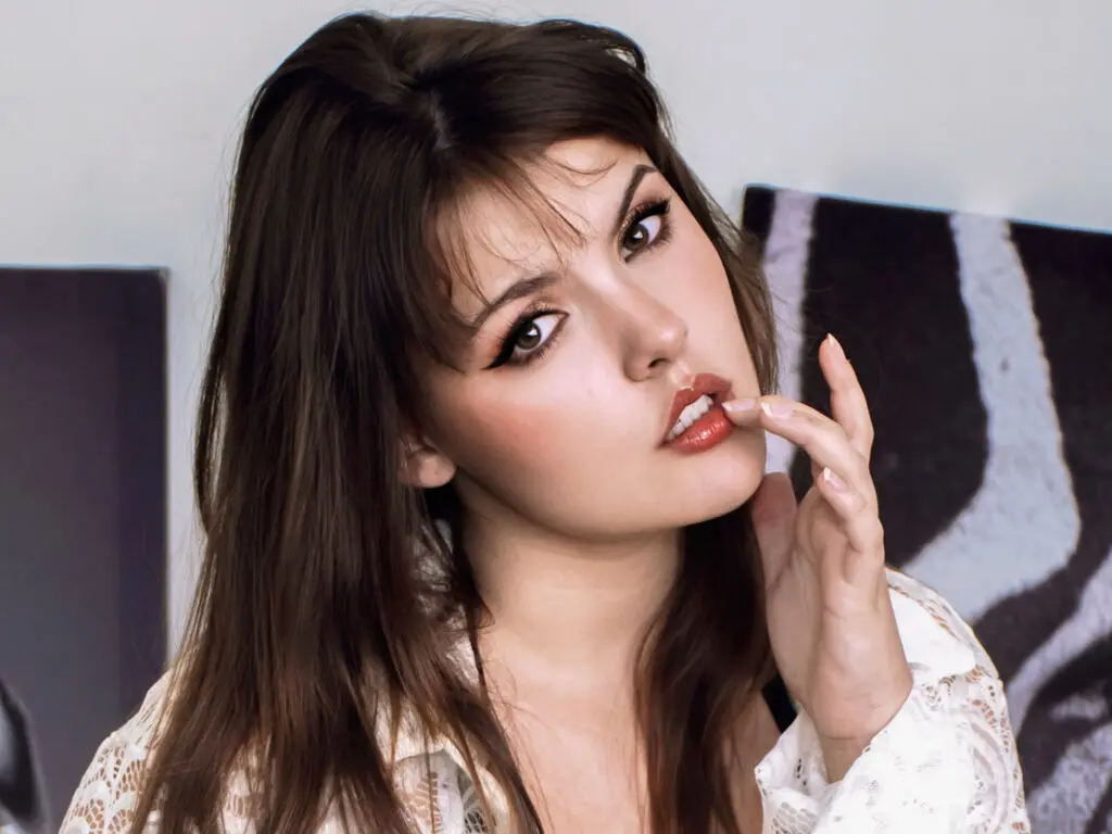 Young woman with brunette hair and winged eyeliner wearing a white lace blouse in a soft, moody indoor setting.
