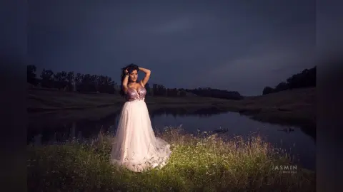 MissyJolie stands in a grassy field by a lake at dusk, wearing a purple lace bustier and a long white tulle skirt.