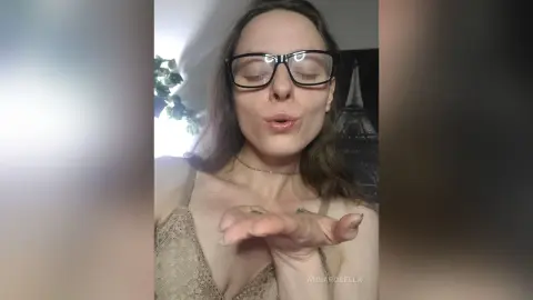 Young woman with glasses in a lace camisole blowing a kiss in a cozy indoor setting.