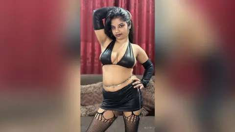 Sejuti poses in a black bikini top, a short skirt, and long gloves with fishnet stockings against a red curtain background.