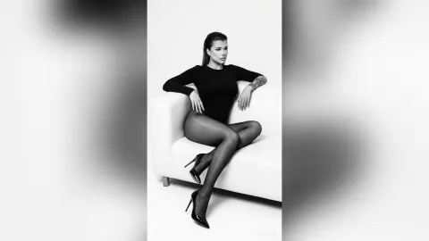 AvaBronX — AvaBronX sits on a white sofa wearing a tight black long-sleeve bodysuit and sheer black tights.
