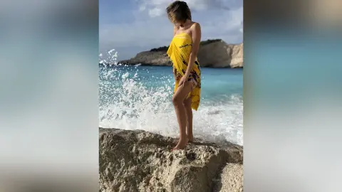 SandraMidnight stands naked on a light-colored rock by the turquoise ocean, wrapped in a bright yellow sarong.