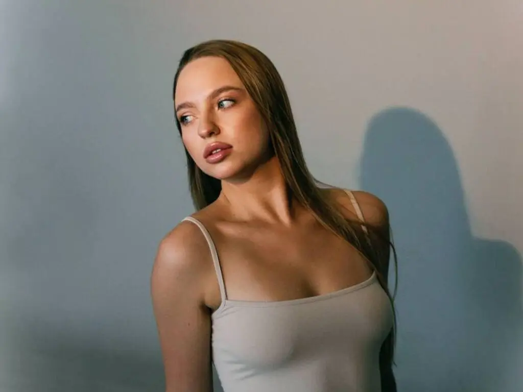 Young woman in a beige camisole tank top posing against a blue wall with soft cinematic lighting.