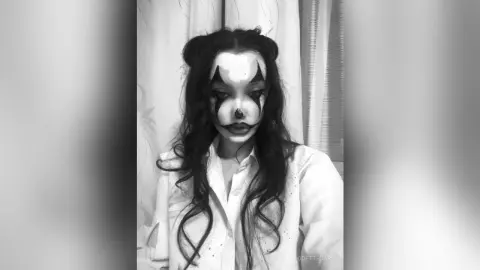 Woman in white shirt with dramatic black clown face paint and wavy hair in a moody black and white portrait.