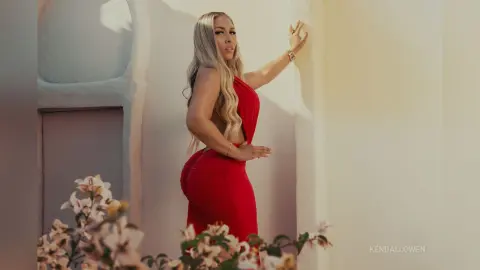 Blonde woman in a glamorous red evening gown posing against a sunlit white wall in a Mediterranean setting.