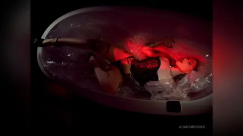 AdaraBrooks lies in a white bathtub lined with plastic, wearing black lace lingerie under moody red lighting.