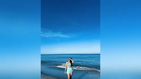 Young woman in a light blue bikini standing on a sunny beach under a clear blue sky.