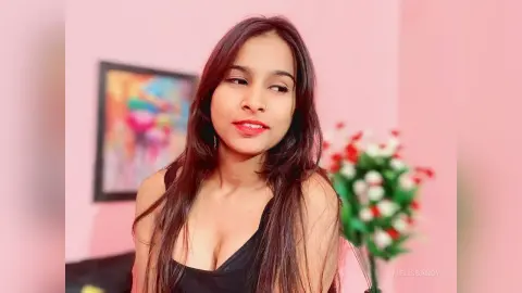 Young woman with long brown hair wearing a black tank top in a bright pink room with floral decor.