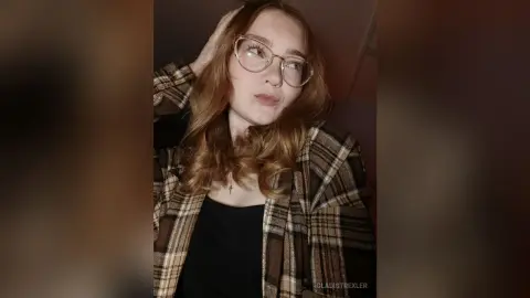 Young woman with glasses wearing a brown plaid flannel shirt in a cozy, moody indoor setting.