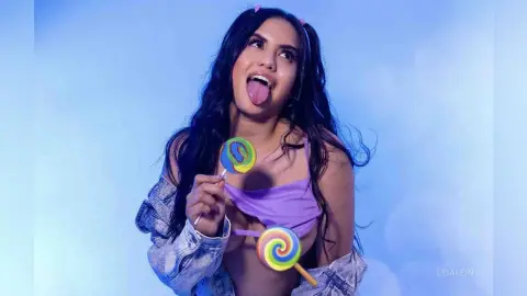 LeiaLein — Playful young woman in a purple top and denim jacket holding colorful swirl lollipops against a blue background.