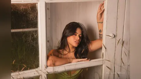 Dreamy portrait of a tattooed woman in a green dress leaning against a white window frame in soft natural light.