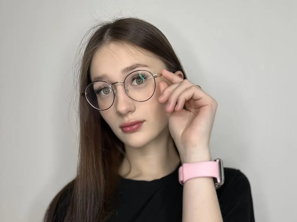 Young woman with long brown hair and round glasses wearing a black shirt in a minimalist indoor setting.